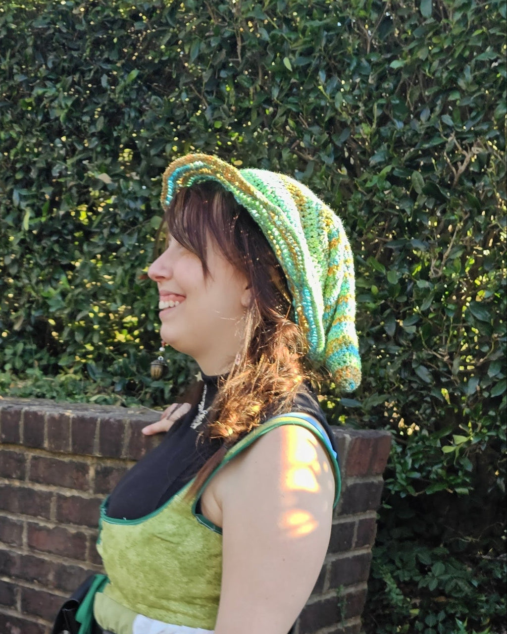 Woman wearing a multi color green, blue, and white crocheted witch hat standing in front of bushes and a brick wall
