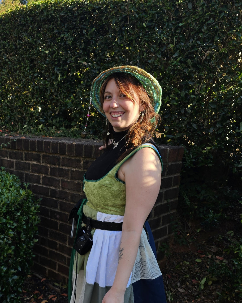 Woman wearing a multi color green, blue, and white crocheted witch hat standing in front of bushes and a brick wall