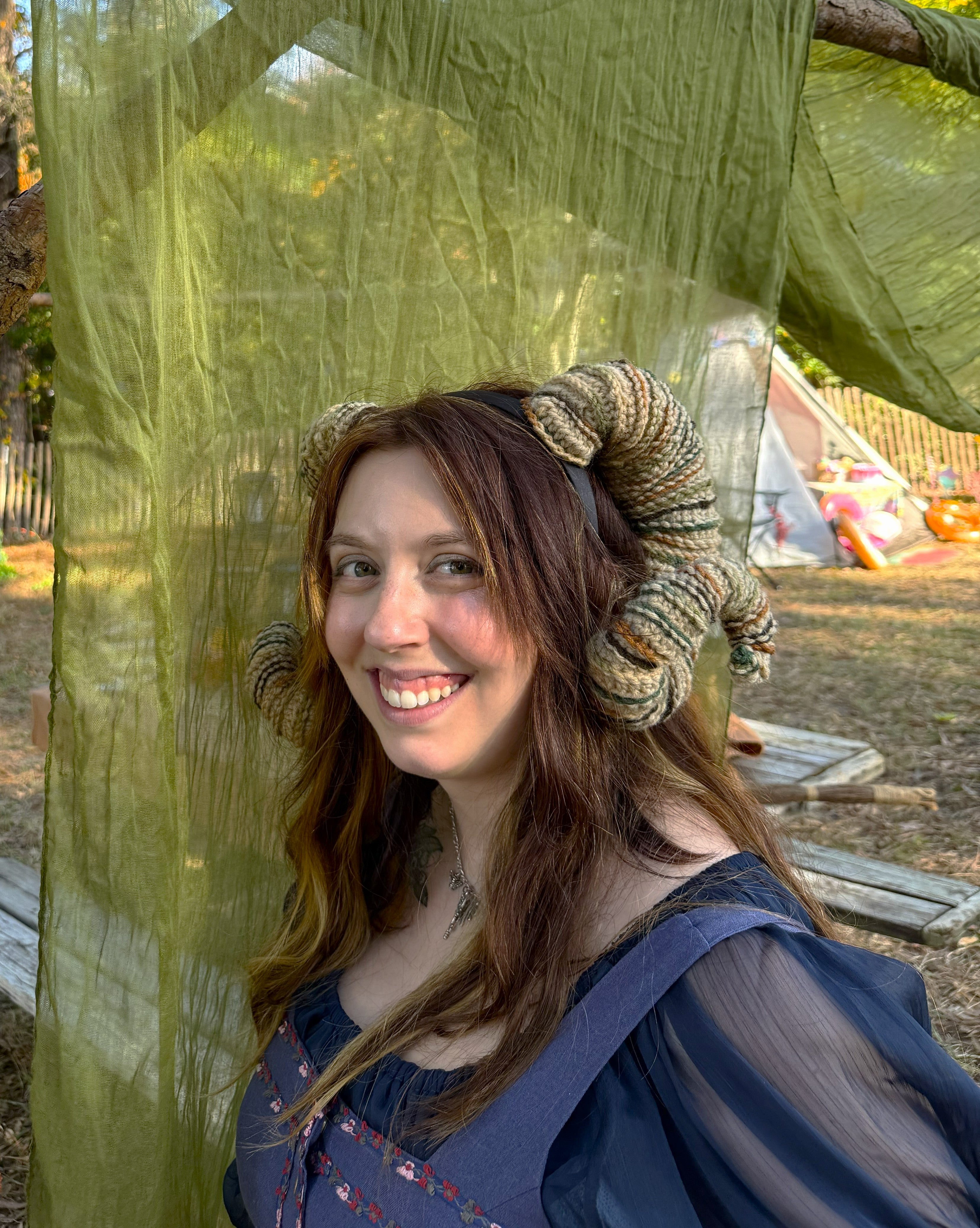 Woman with ram horns and blue dress standing in front of green fabric